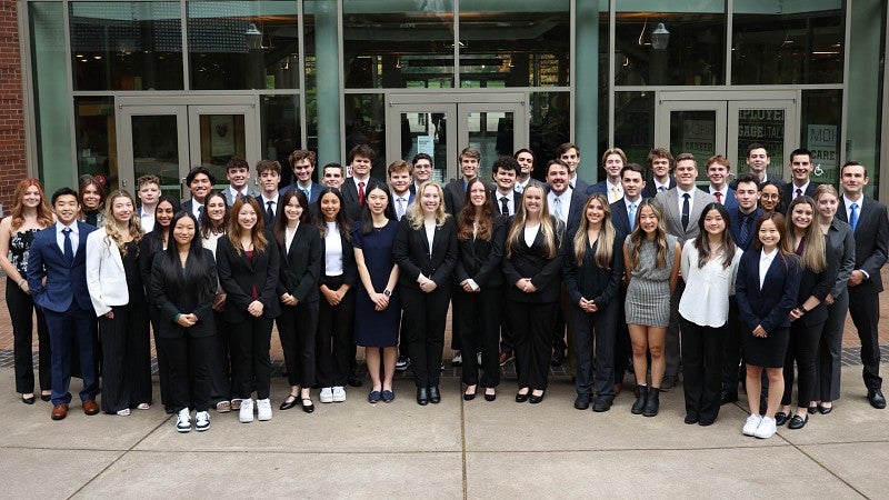 A group of honors students stands gathered for a photo in front of the north entrance to the Lillis Business Complex