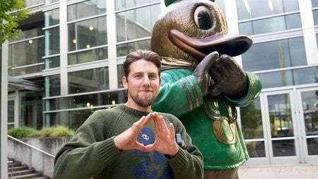 Lundquist College alumnus Orion Falvey, standing beside the Duck mascot statue outside the Ford Alumni Center, makes the "O" symbol with his hands