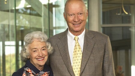 Patricia Peterson (left) and husband Ron Peterson (right) stand smiling together in the atrium of the Lillis Business Complex