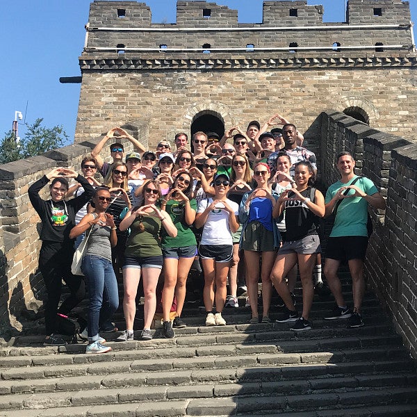 Large group of students standing on the steps of the Great Wall of China, looking at the camera, making an O shape with their hands.