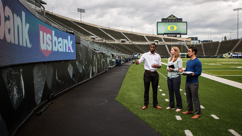 Three students standing on the field of Autzen Stadium looking at a sponsor banner